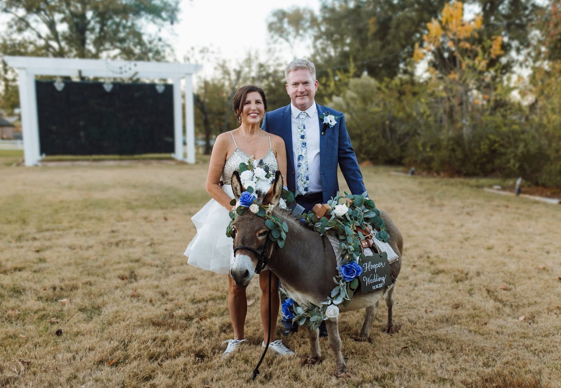 Bride and groom pose with donkey decorated for a wedding. They are outside near a floral backdrop.
