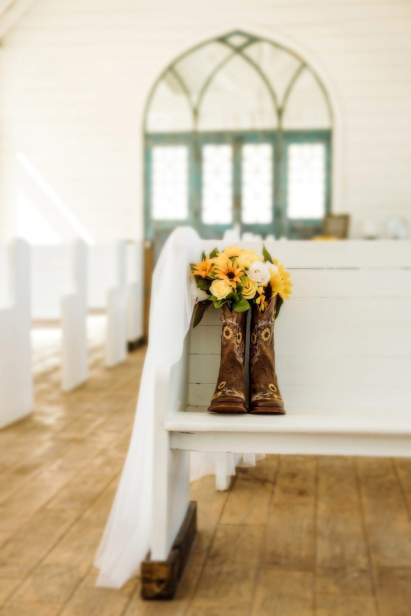 Brown cowboy boots and yellow flowers on a white bench in a church, with a veil draped over it.
