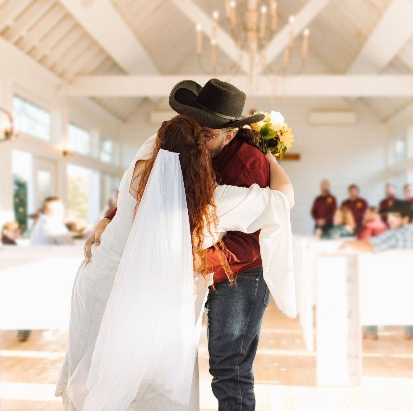 Bride and person in a cowboy hat embracing, inside a bright room with tables and wedding party.