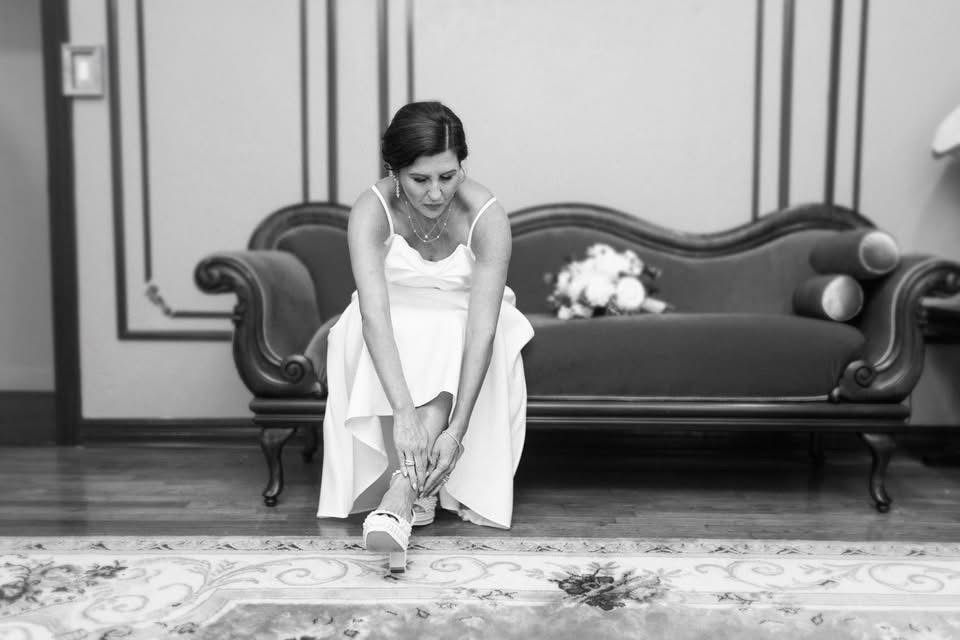 Bride puts on her shoe, sitting on a vintage couch. White dress, bouquet on couch, ornate room.