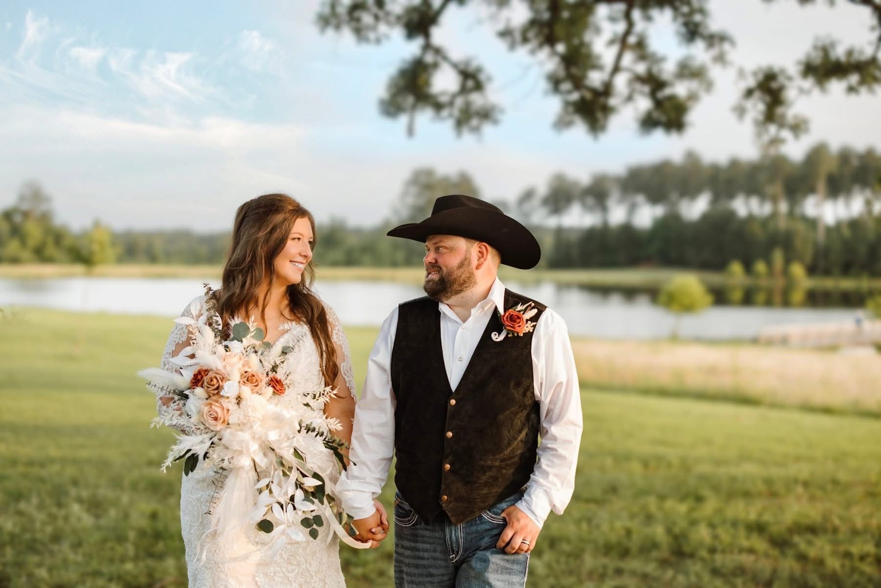 Bride and groom holding hands, smiling at each other. They're in a field near a lake. He wears a cowboy hat, she holds a bouquet.