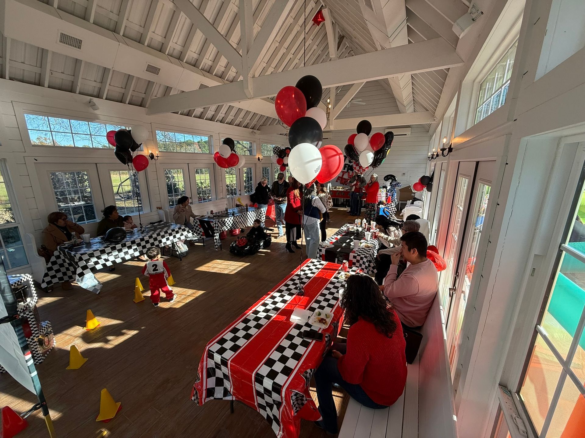 Party with race car theme; guests sitting at checkered tables, balloons in red, white, and black.