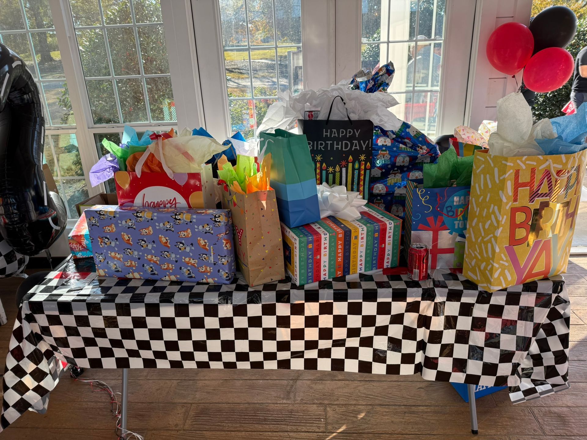Gifts on a table with a black and white checkered tablecloth. Balloons and window in background.