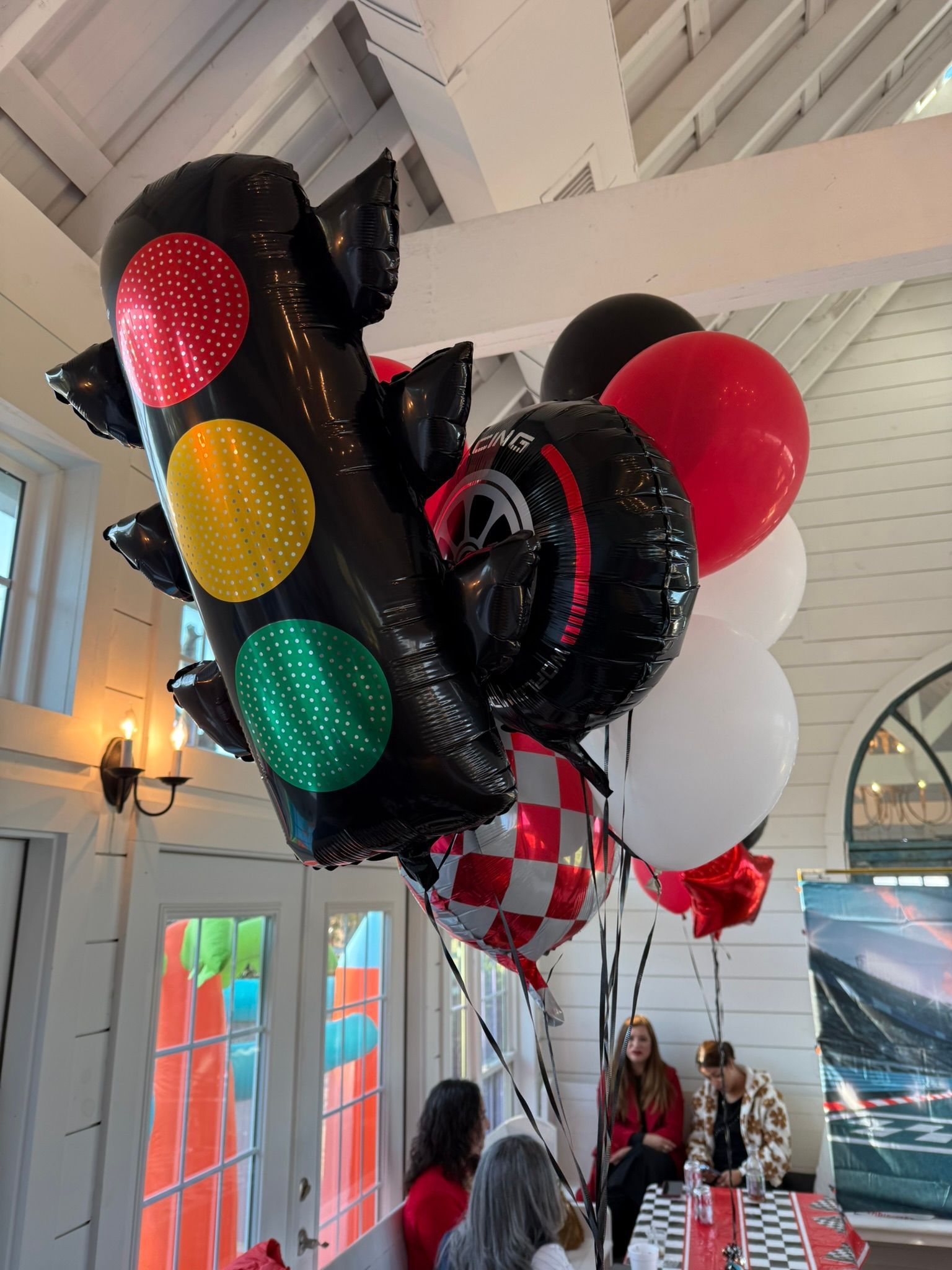 Race car themed balloons: traffic light, tire, red/checkered balloons; people in setting.