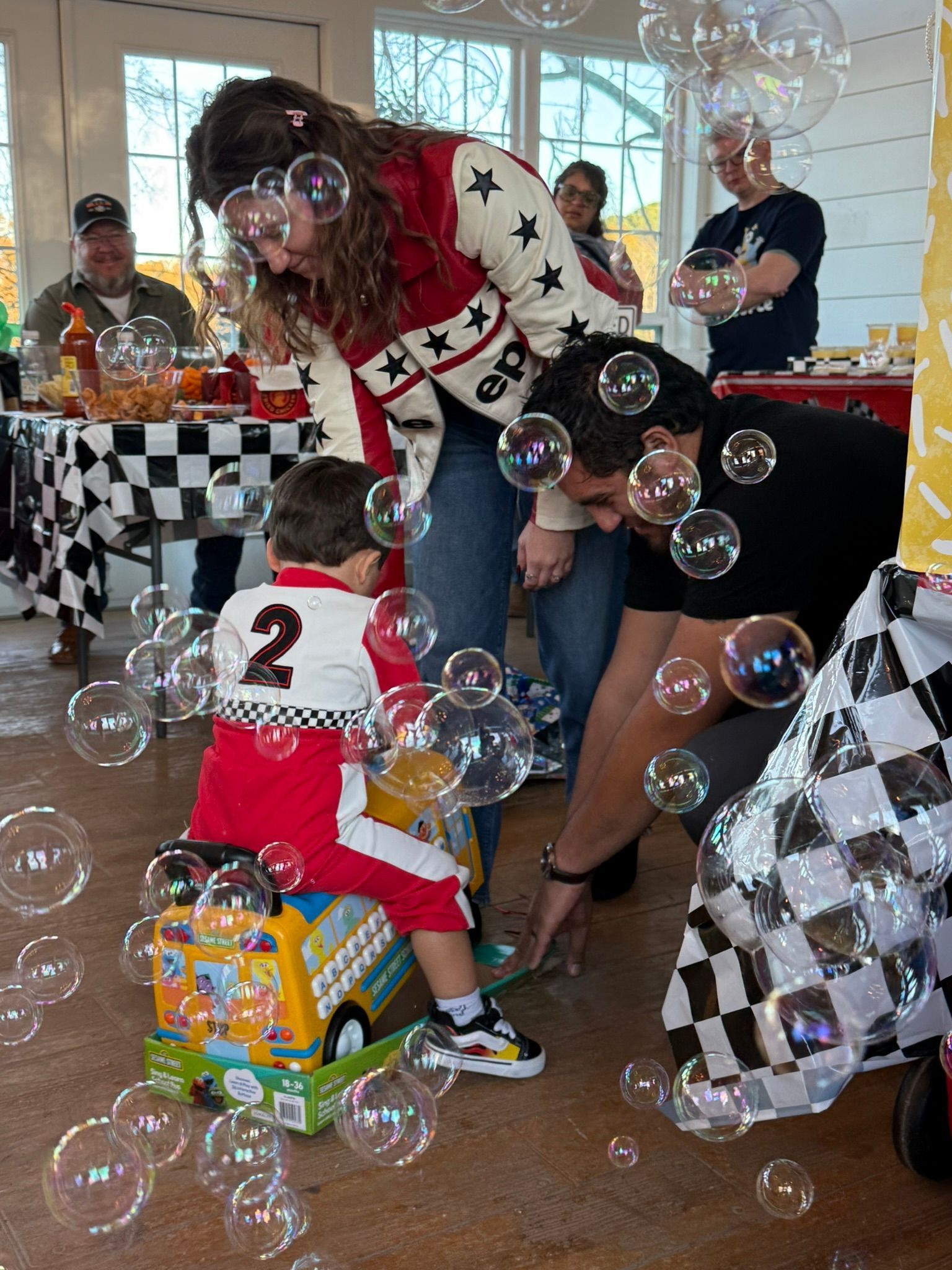 Family blows bubbles. Child in racing suit sits on toy bus. People at table, checkered tablecloth.