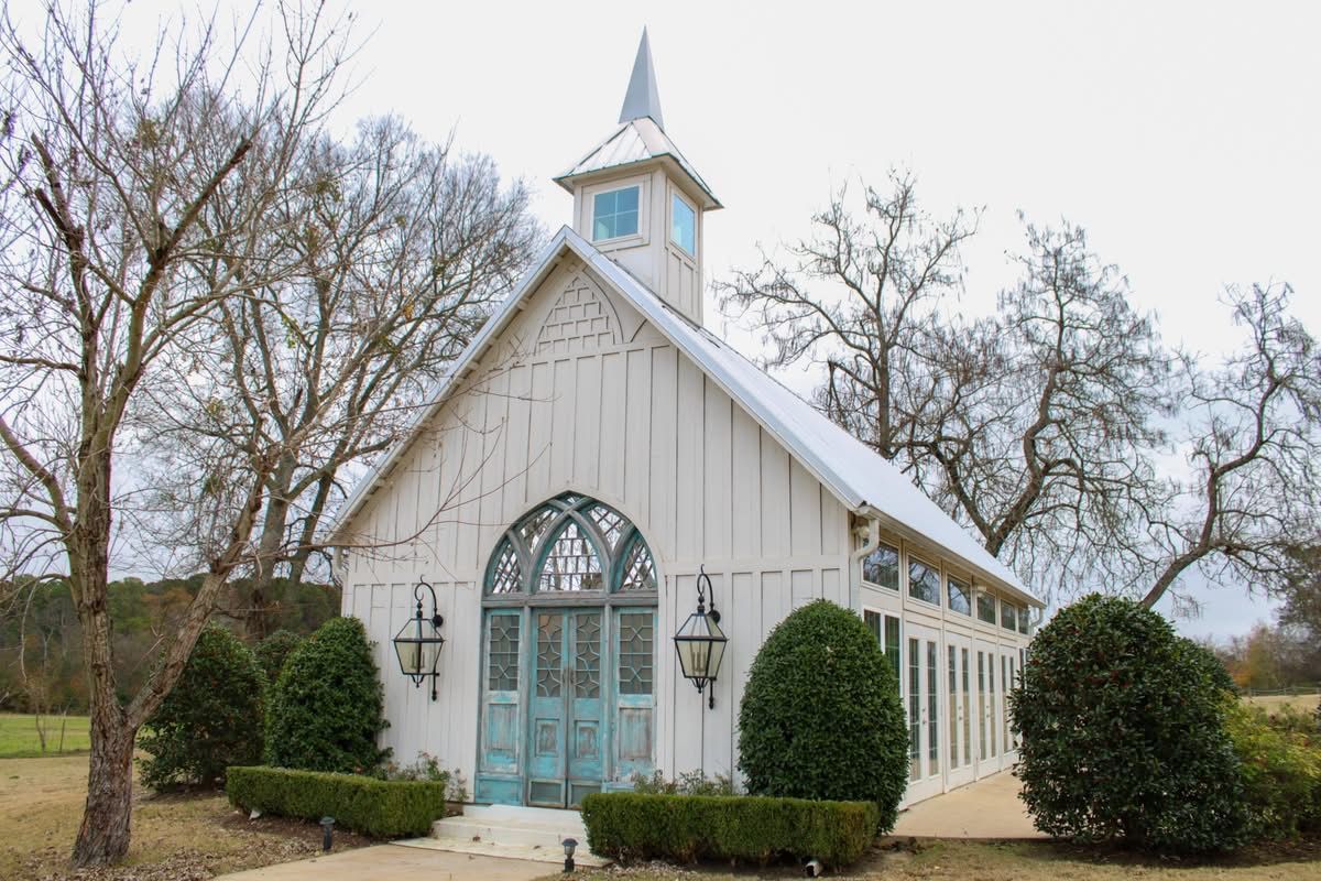 White chapel with blue doors, steeple, and arched window, surrounded by trees and bushes.