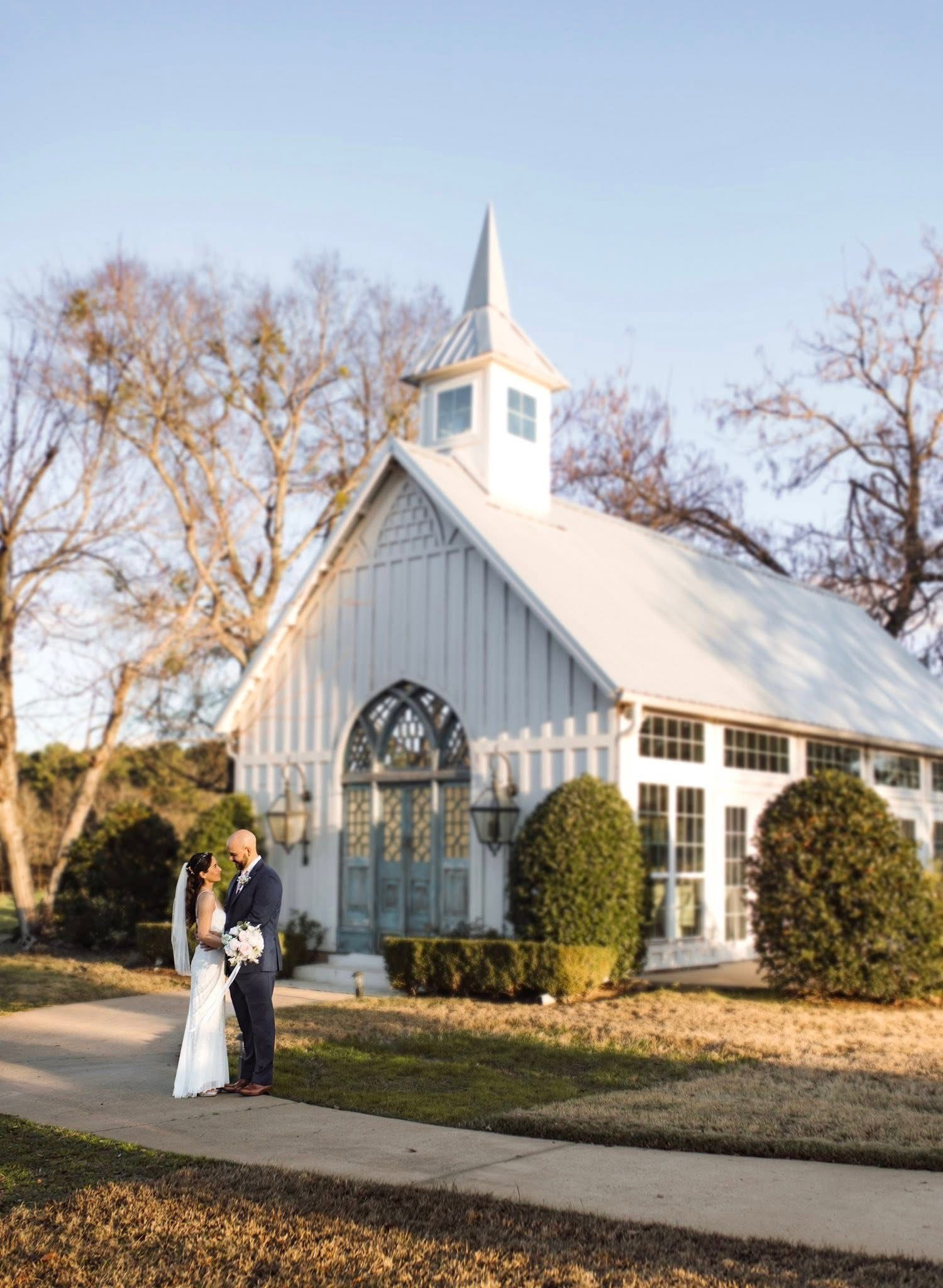 Couple embraces outside a white chapel with blue doors; sunlight and trees.