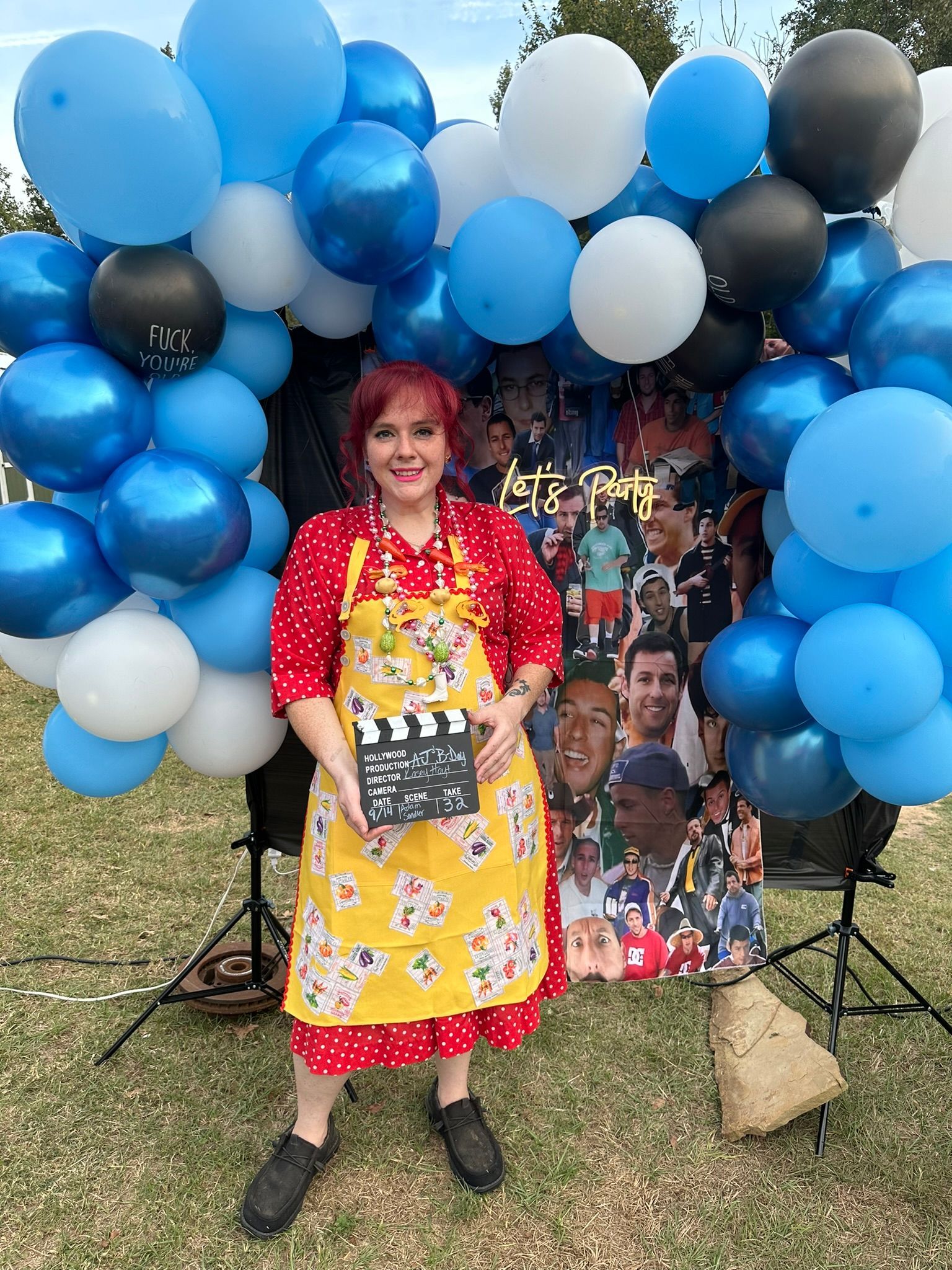 Woman in red dress and apron holds a clapperboard in front of a balloon arch and sports banner.
