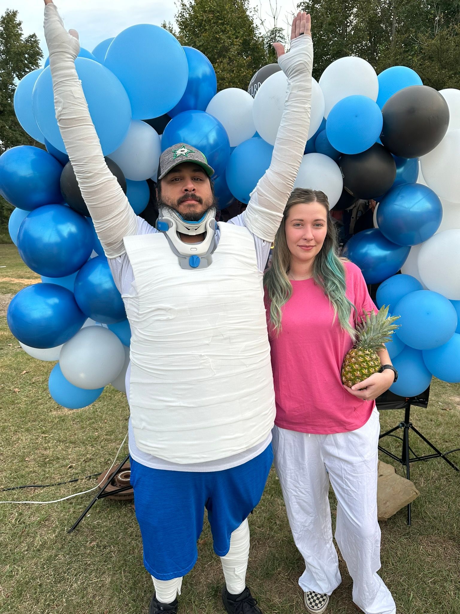 Man in neck brace and casts with arms raised, woman holding pineapple, blue balloons. Outdoor setting.