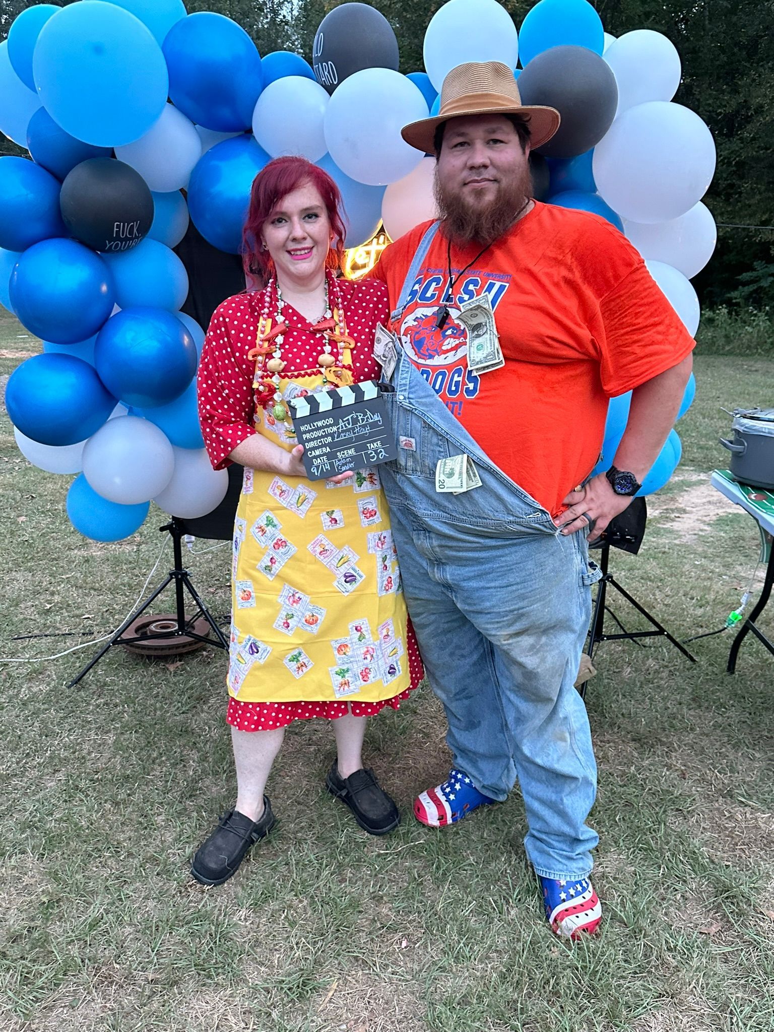 Couple in costumes pose with film clapper in front of balloons; one wears overalls, hat, and red shirt.