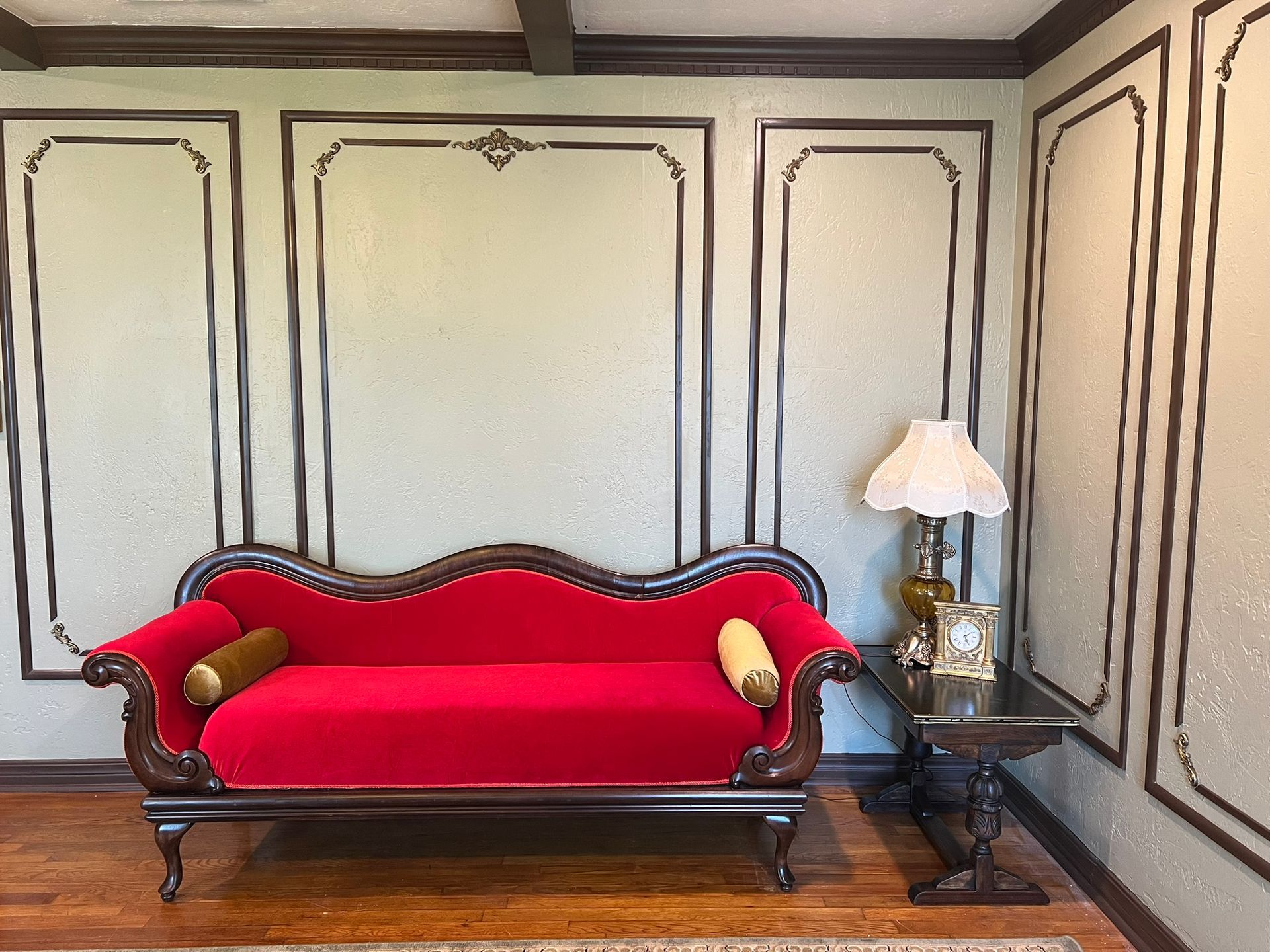 Red velvet sofa in a room with ornate wall panels, a side table with a lamp, and a wooden beam ceiling.