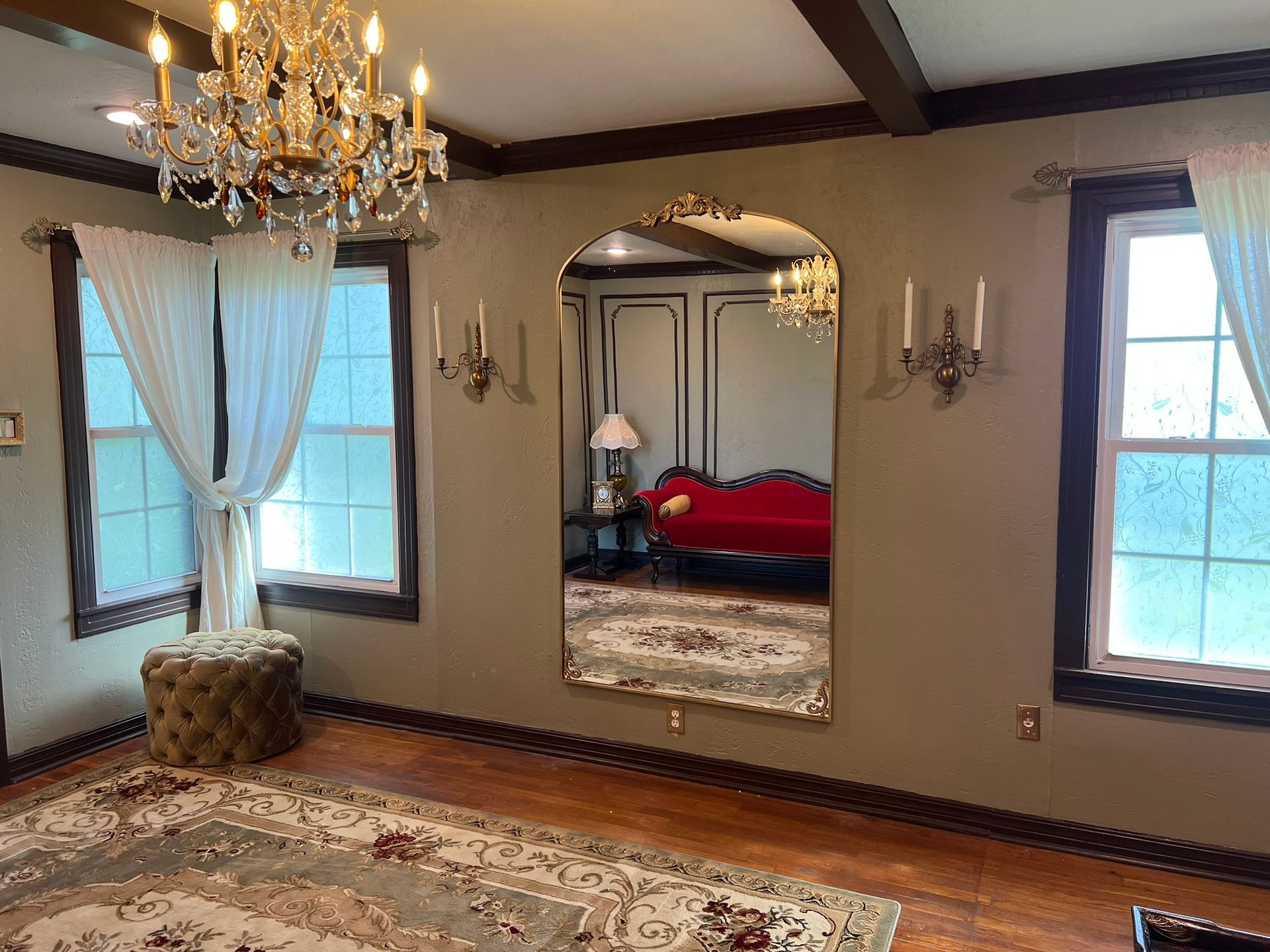 Elegant room with chandelier, large mirror reflecting a red sofa, windows, and patterned rug.