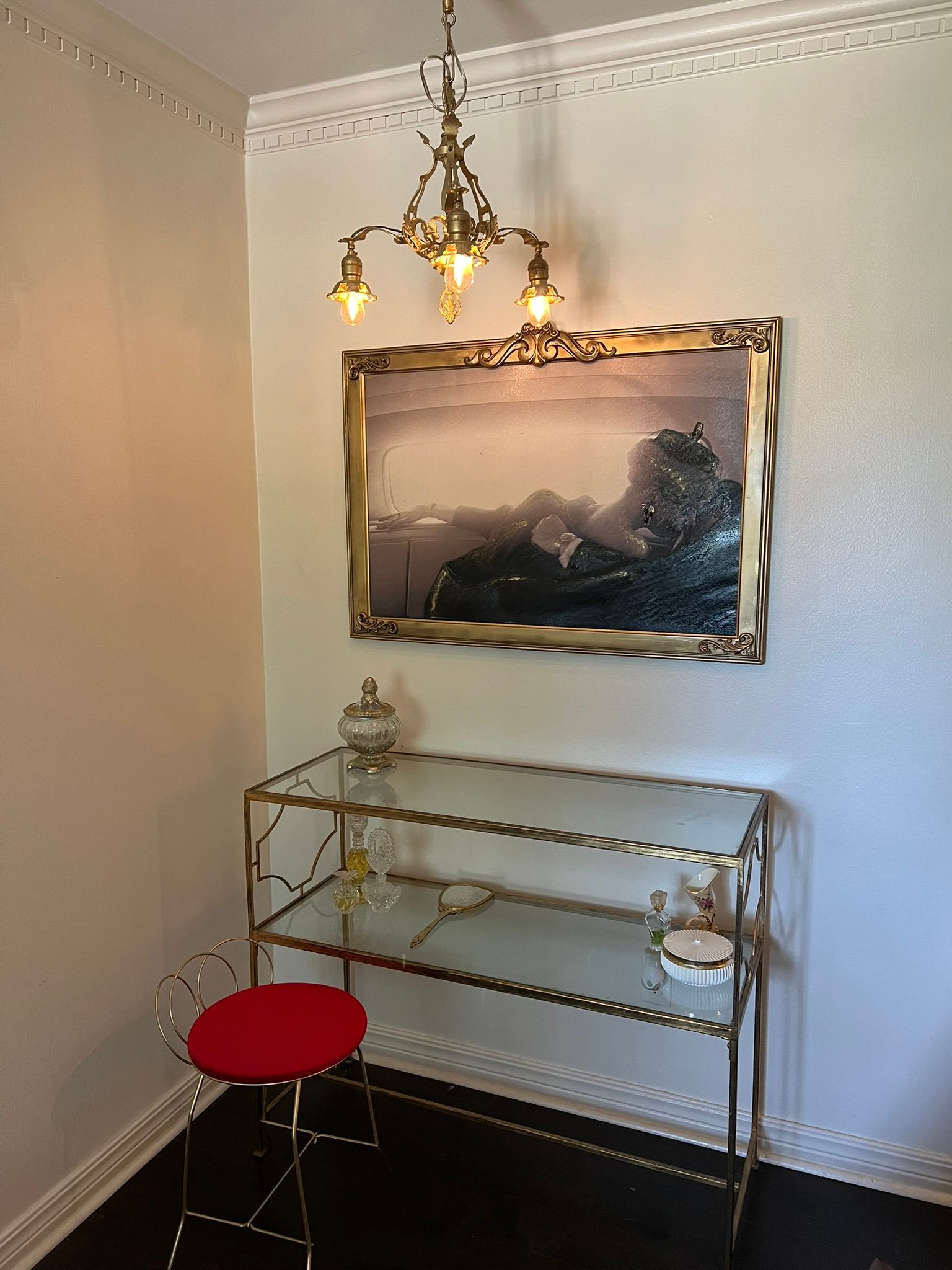 A small room with a painting, glass shelf, red stool, and chandelier.
