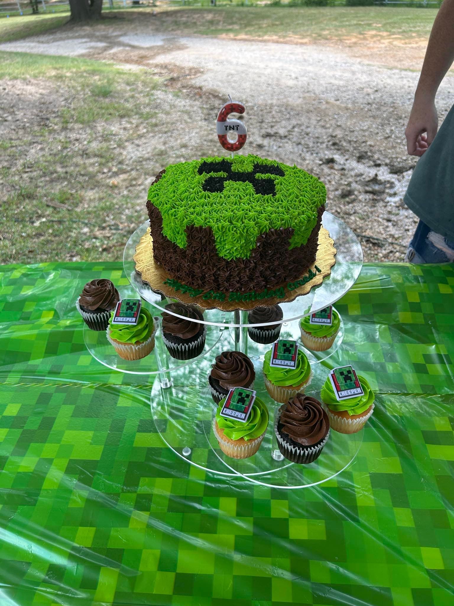 Minecraft-themed birthday cake and cupcakes on a transparent tiered stand, green and brown frosting, 