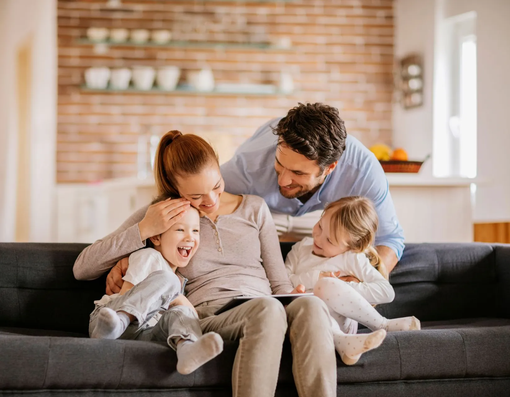 Family of four relaxing together on a couch in a bright living room, smiling and hugging.