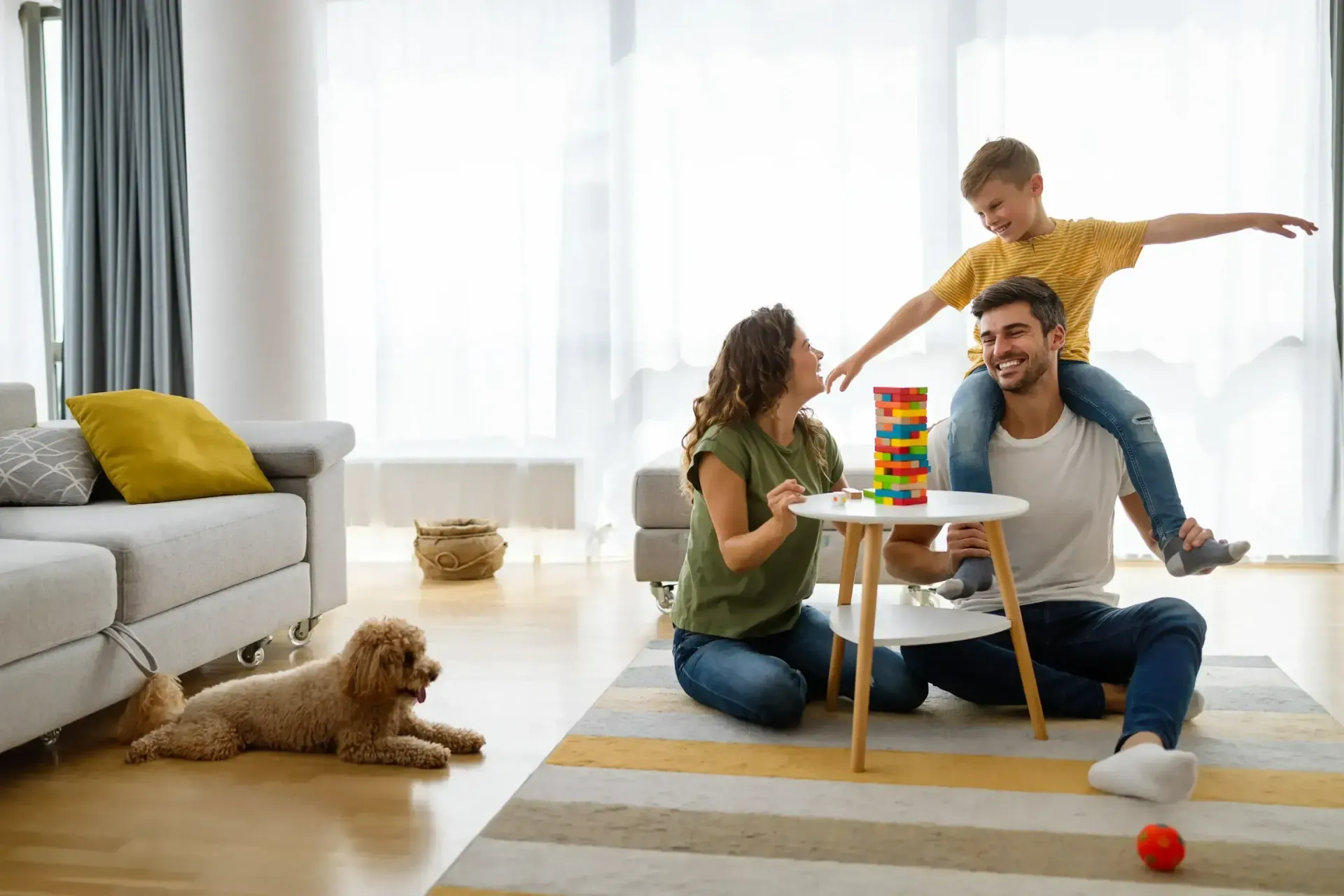 Family playing a stacking game in a bright living room with a dog resting nearby.