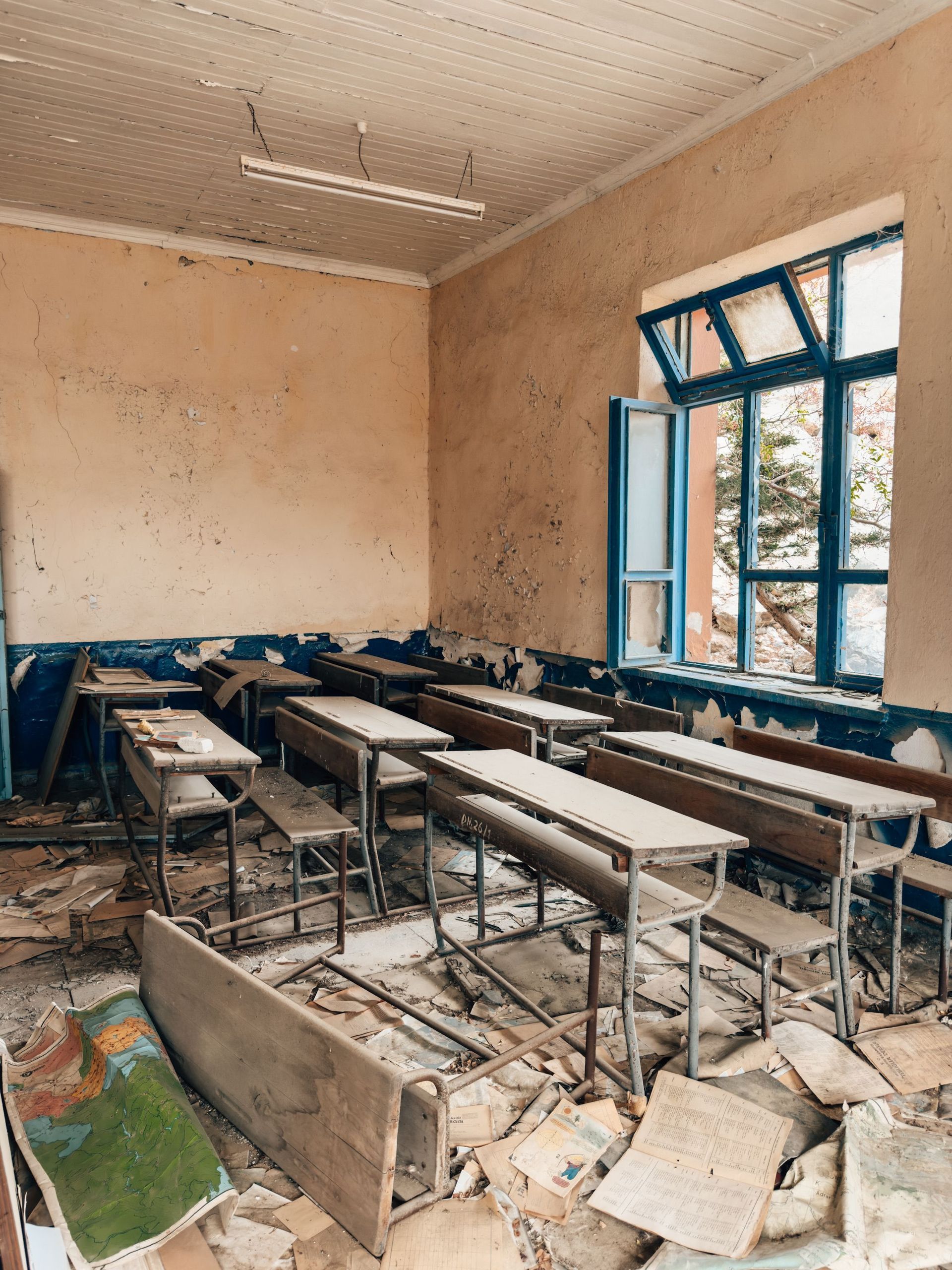 Damaged classroom with broken desks, debris, and open blue windows letting in daylight