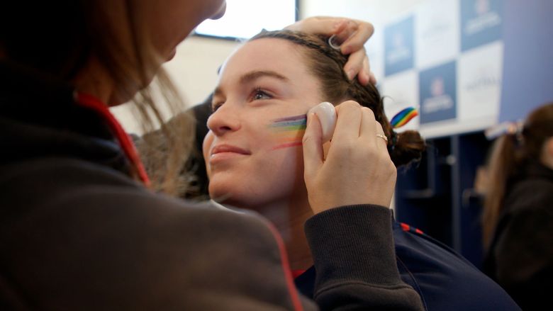 Person paints rainbow stripes on another person's cheek at a crowd event.