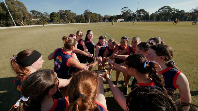 Women in black and red sports uniforms huddle together on a grassy field, hands in for a team cheer.