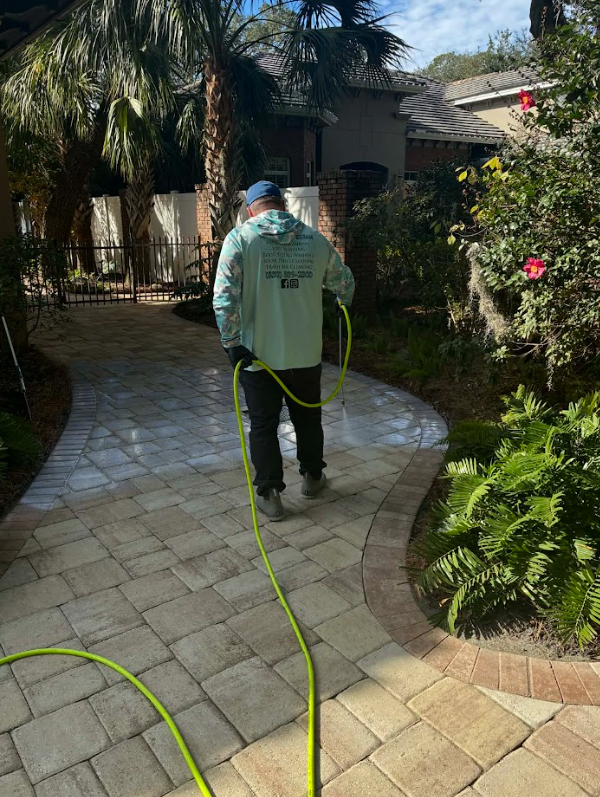 A person in a blue hat sprays a paver path with a green hose, near bushes and a house.