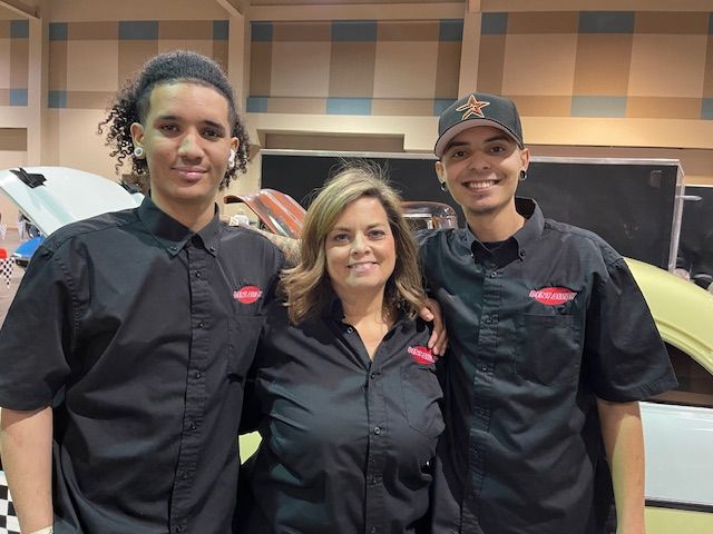 Three people in black shirts pose in front of a car at an event.