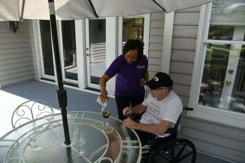 Woman pours liquid into a glass for a person in a wheelchair on a patio, under an umbrella.