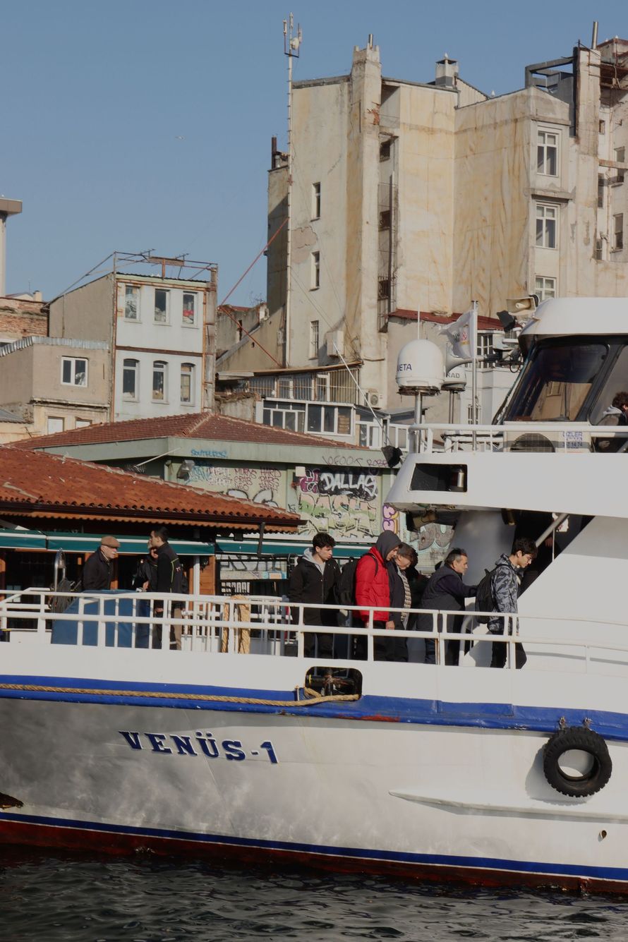 Crowded white boat with blue trim docked by old waterfront buildings and a small market