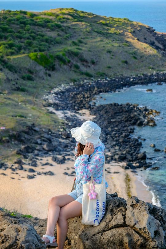 Person in a white sunhat sits on a rocky cliff overlooking a curving blue bay and green coastline.