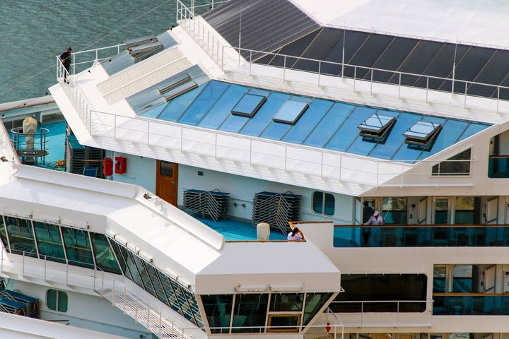 Cruise ship deck with blue rooftop panels and passengers on a white walkway by the sea