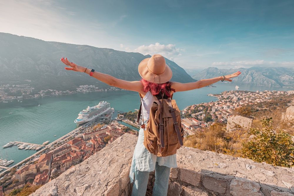 Traveler with backpack and hat, arms outstretched on a stone overlook above a coastal mountain town and blue bay