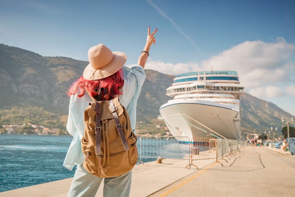Traveler with backpack and hat waves toward a large cruise ship at a sunny seaside port.