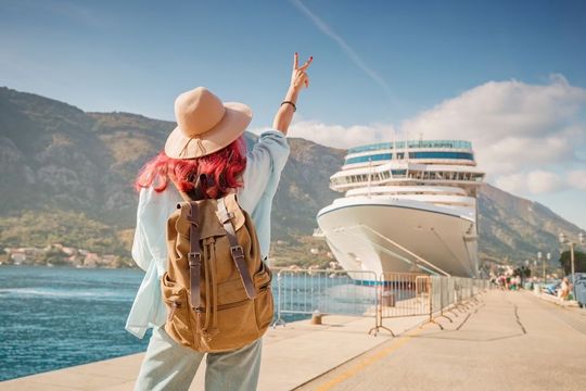 Traveler with backpack and hat waves toward a large cruise ship at a sunny seaside port.