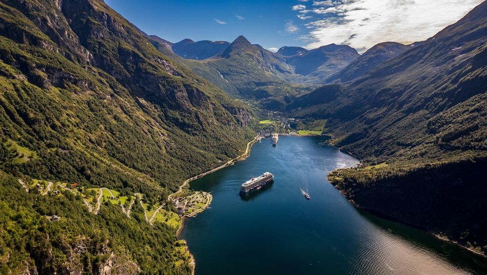 Aerial view of a fjord with a cruise ship, steep green mountains, and a small village under a cloudy sky