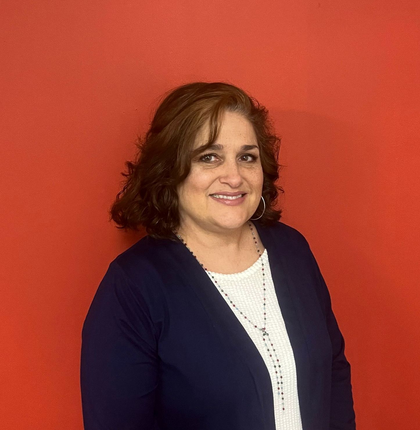 A woman in a blue cardigan and white shirt is smiling in front of a red wall.