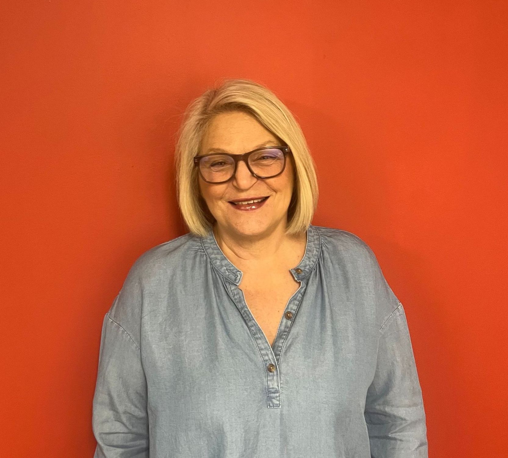 A woman wearing glasses and a blue shirt is smiling in front of a red wall.