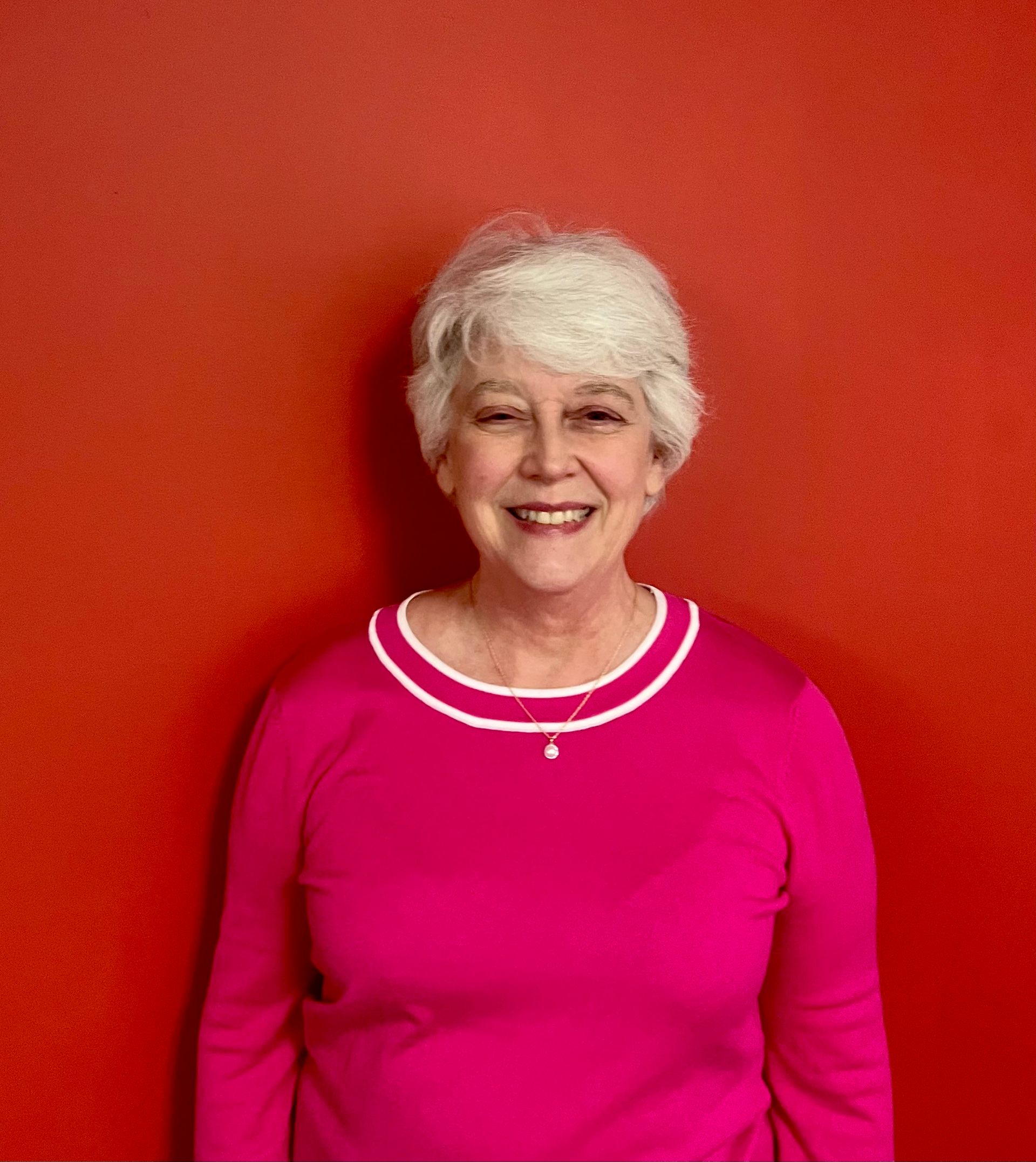 A woman in a pink shirt is smiling in front of a red wall.