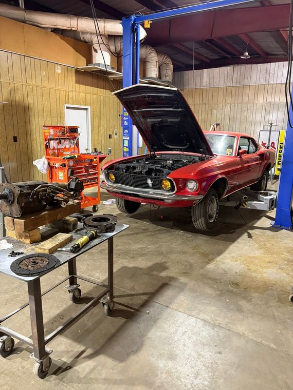 Red classic car in a garage with hood open, lifted on a blue car lift, with tools on a worktable.