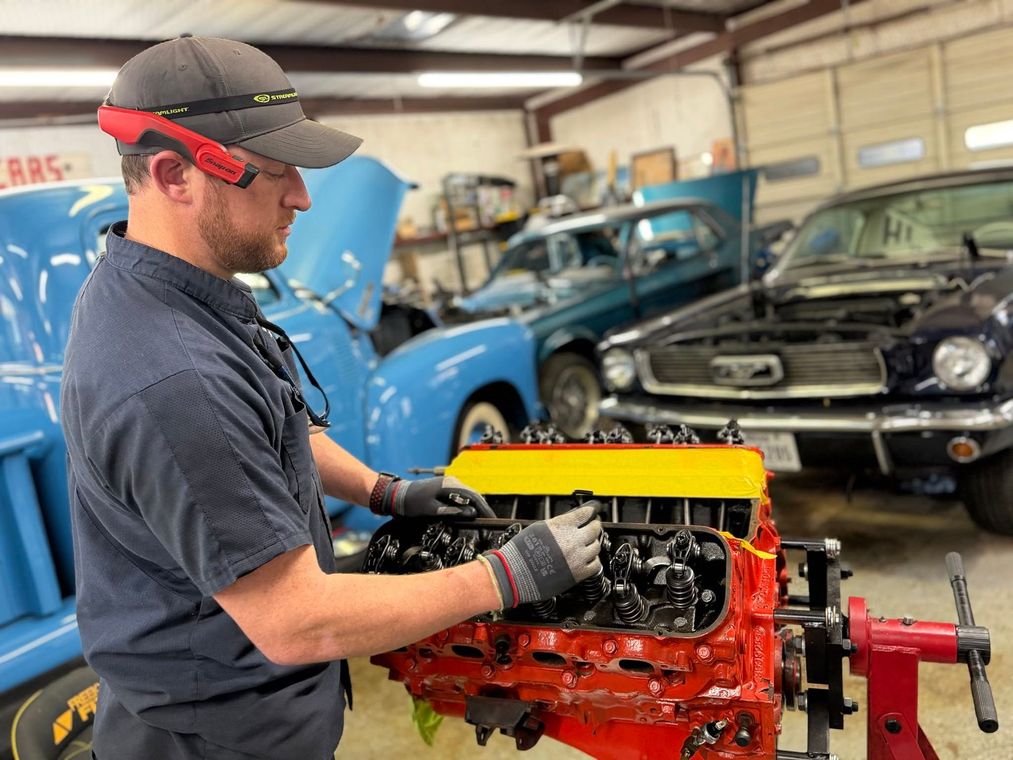 Mechanic working on a red engine in a garage with classic cars in the background