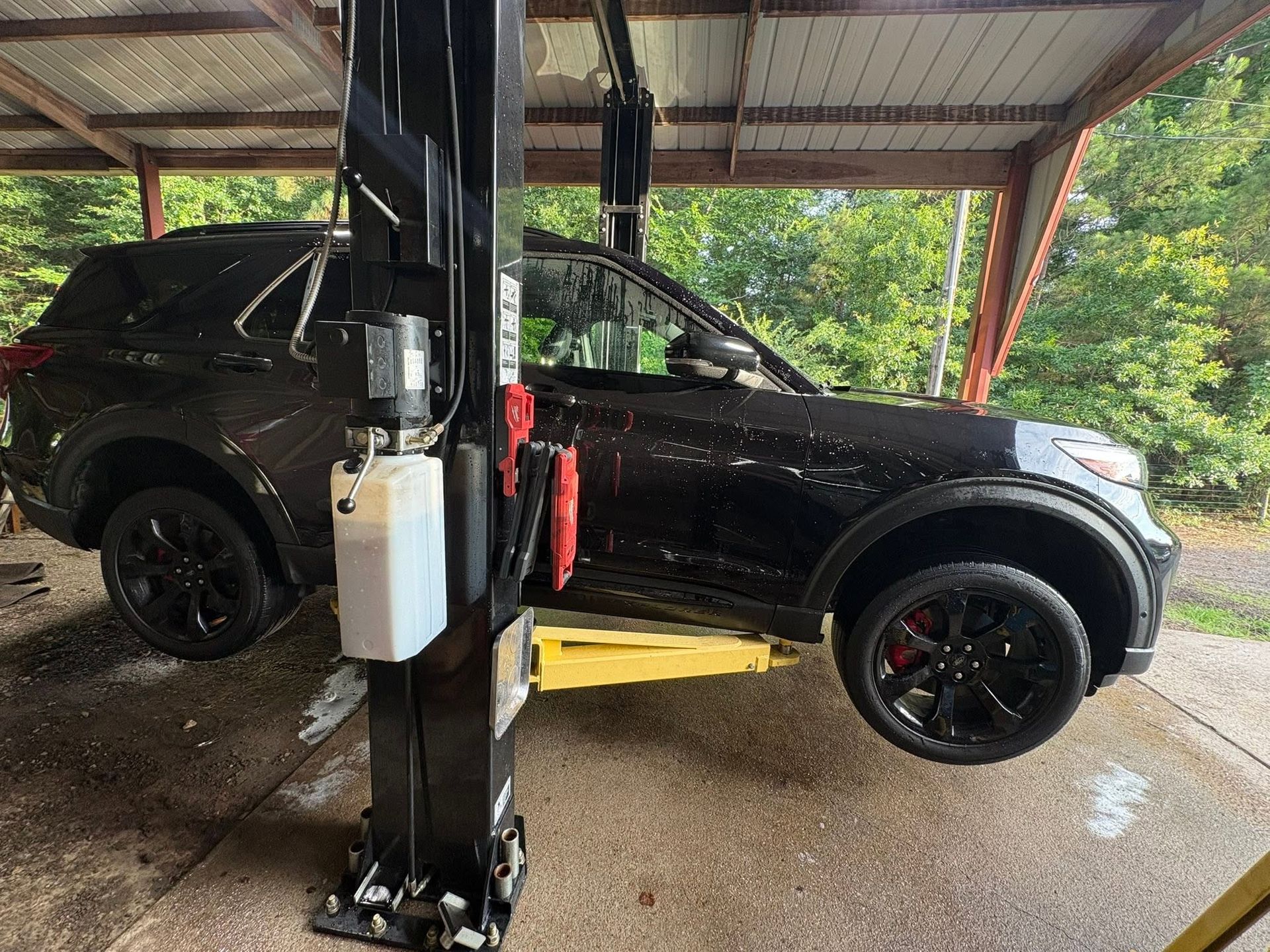 Black SUV on a yellow lift in a wet carport, with a column and trees visible outside.