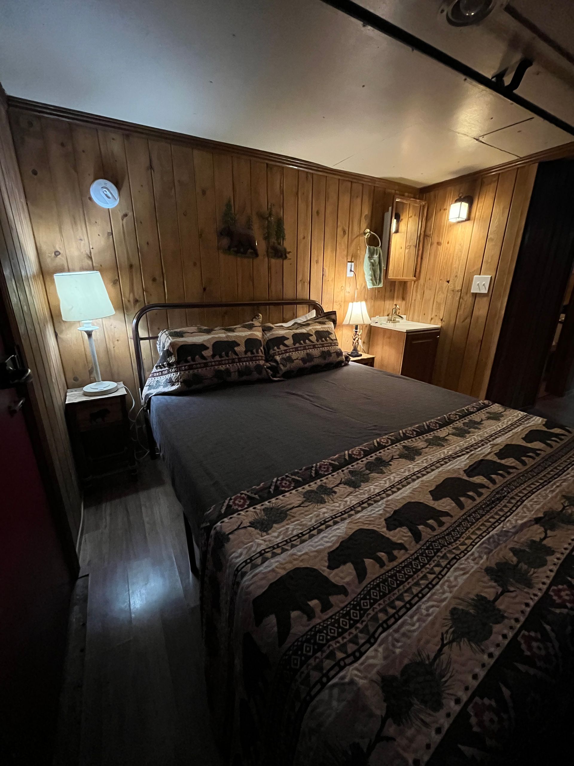 A bedroom with a bed , nightstand , lamp and wooden walls.