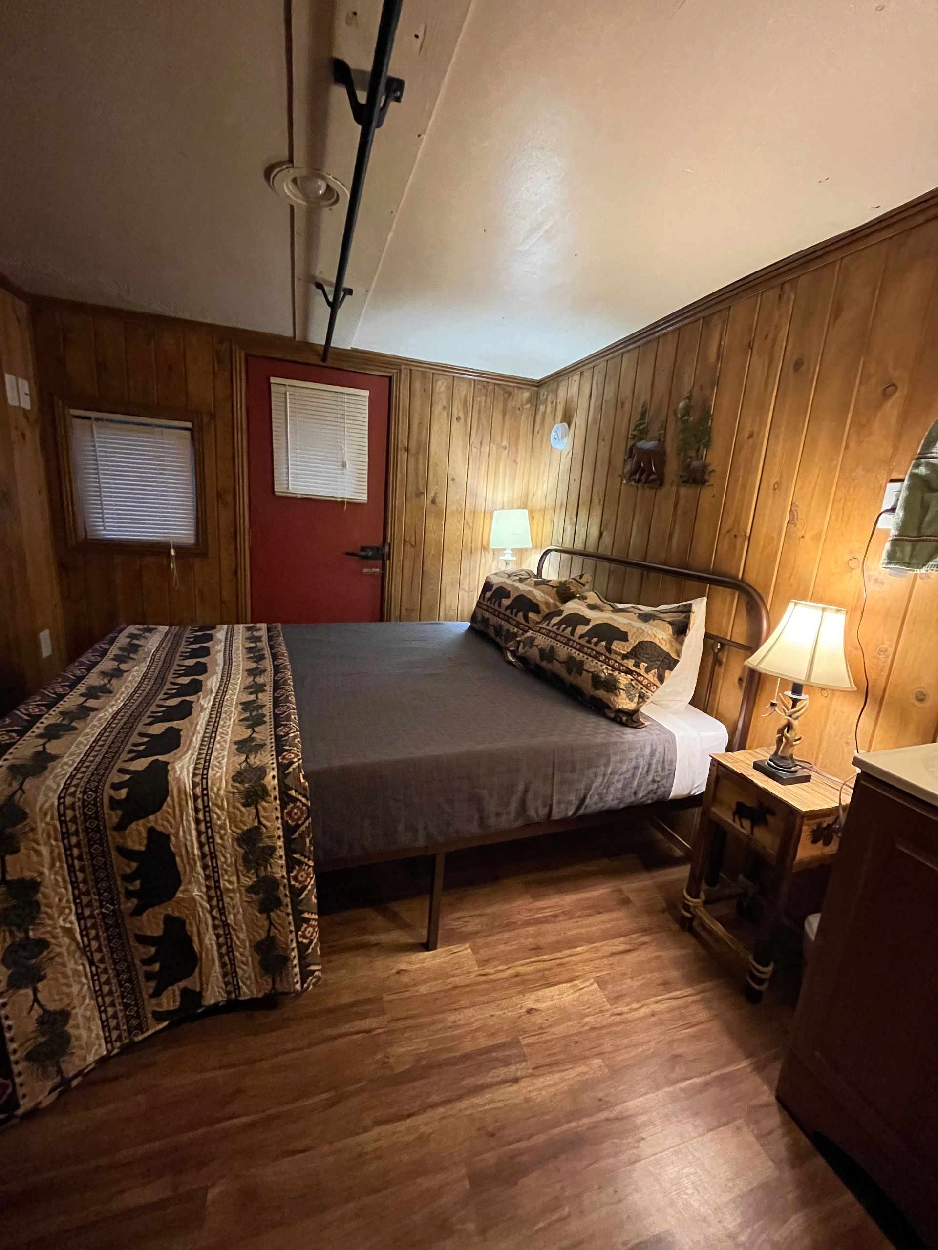 A bedroom with a bed , nightstand , lamp and wooden walls.
