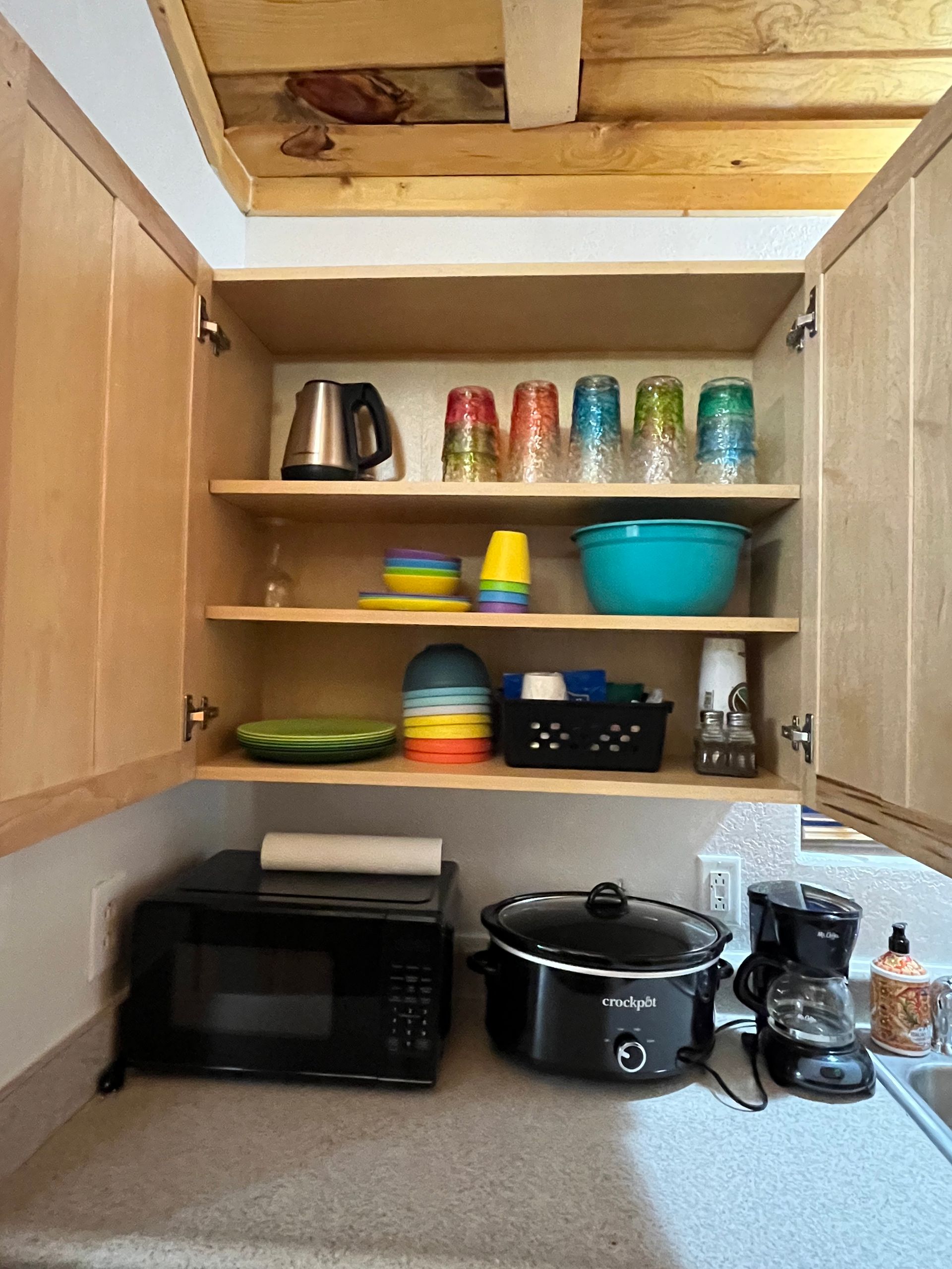 A kitchen with a microwave , crock pot , bowls , and glasses on the shelves.