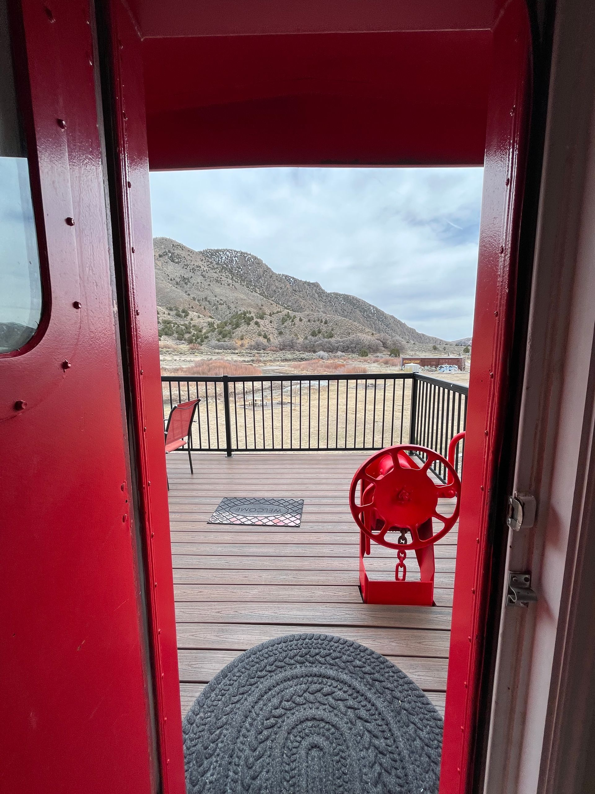 A red door is open to a balcony with a red chair and mountains in the background.