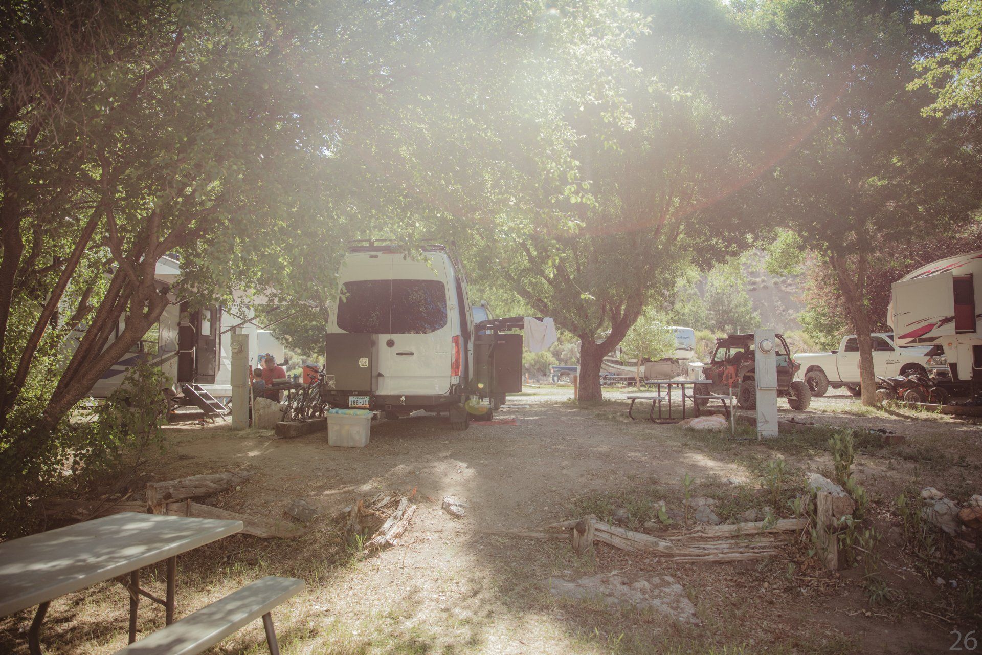 A white van is parked in a campground surrounded by trees.