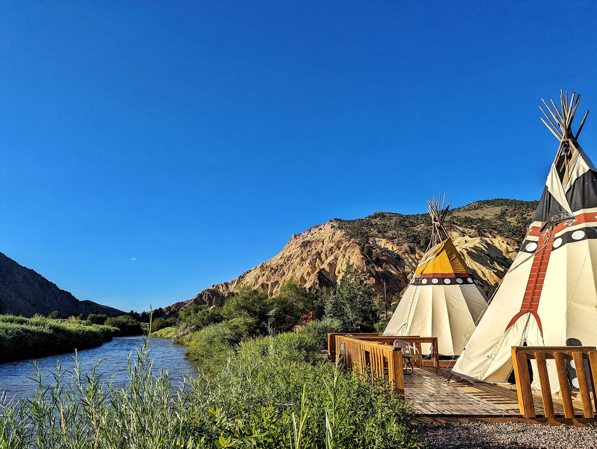 A couple of teepees sitting next to a river with mountains in the background.