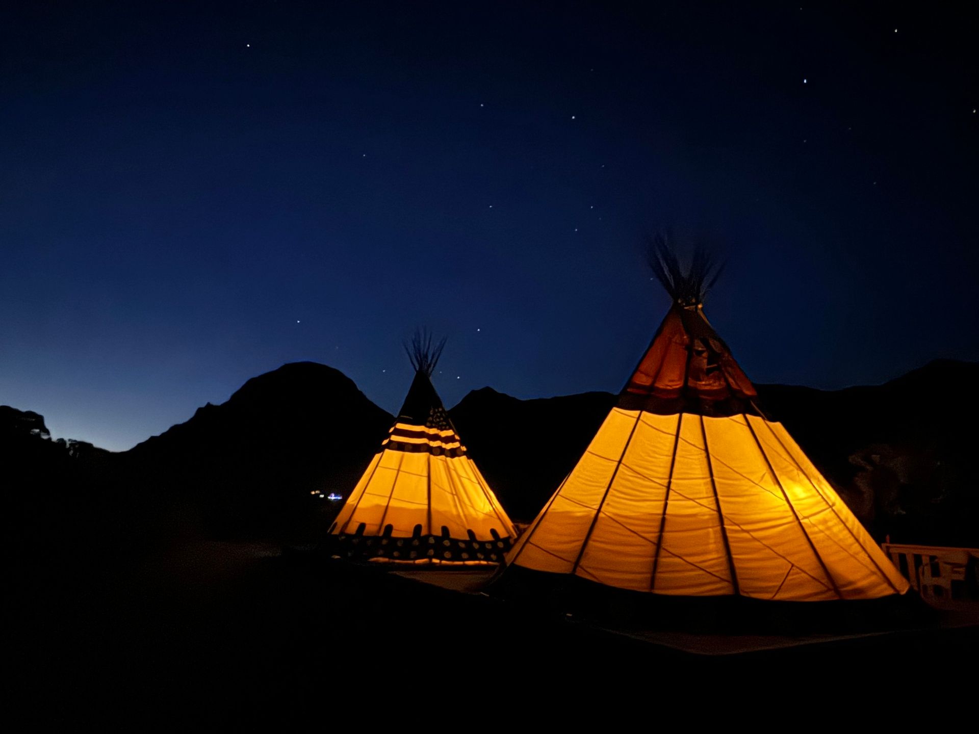 Two teepees are lit up at night with mountains in the background