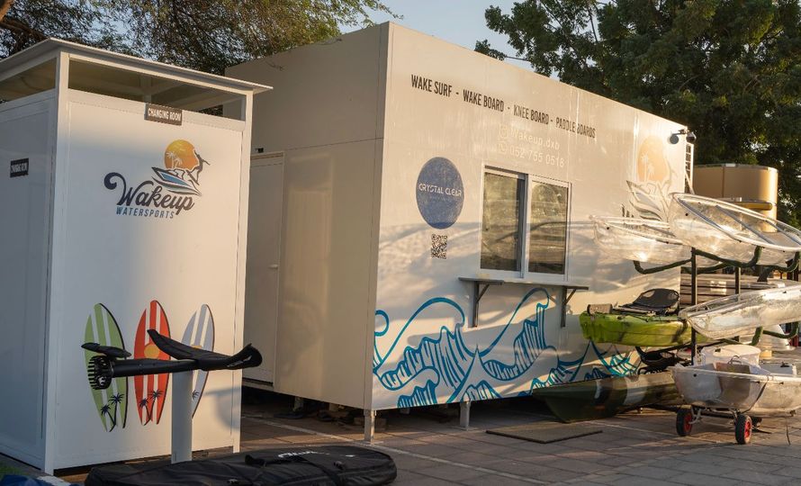 White mobile unit with ocean-themed mural and signage, beside a rack of beach gear outdoors.