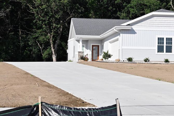 Concrete driveway leading to a white house with a gray roof and front door, surrounded by dirt and trees