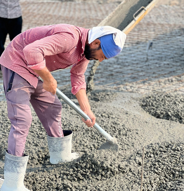 Worker shoveling wet cement at a construction site, wearing a pink shirt, boots, and a head covering