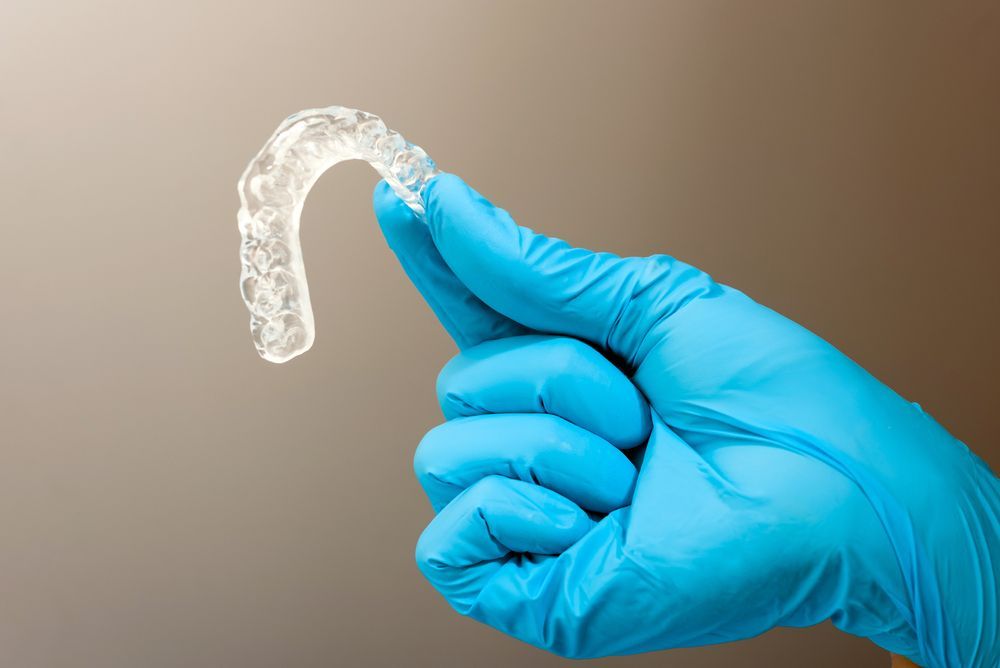 A Hand in a Blue Medical Glove Holds Up a Clear Plastic Orthodontic Aligner — Mareeba Dental Centre in Mareeba, QLD